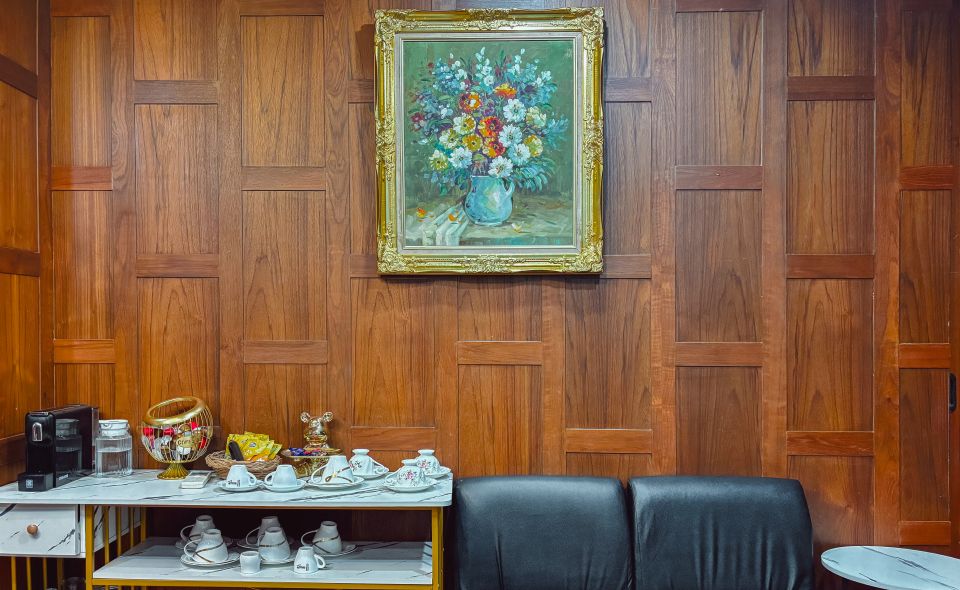 A gilt-framed painting of flowers on walnut panelling, above a black leather sofa and a marble-topped stand with cups and a coffee machine.