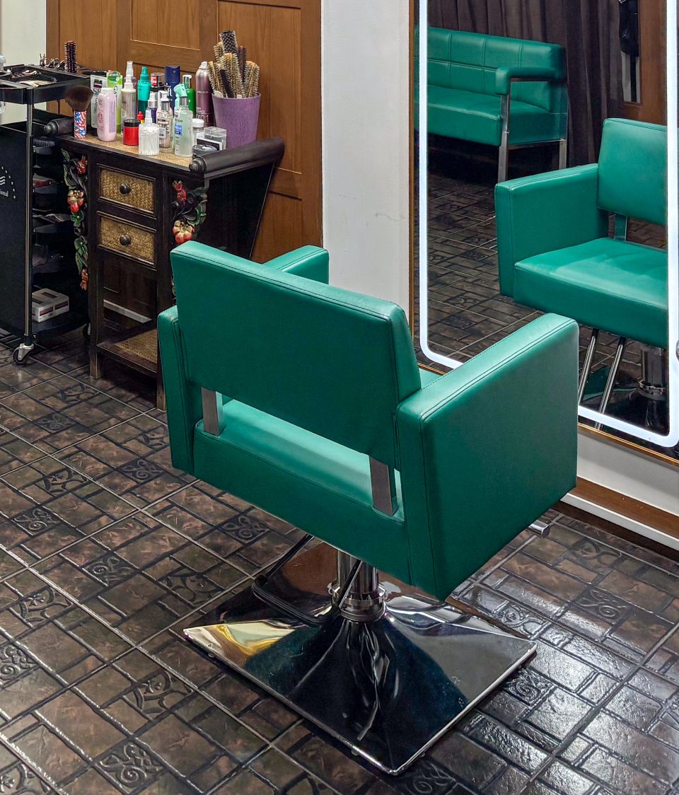 A teal styling chair on a dark patterned tile floor, before a tall mirror lit around its edge that reflects a teal sofa and a second chair.