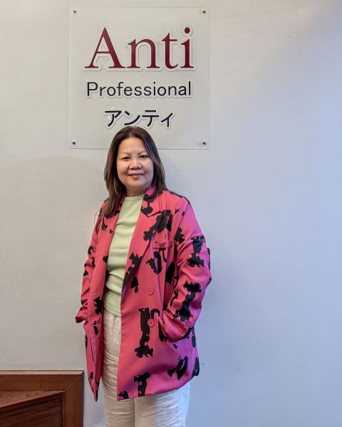 Khun Nuch, smiling, in a pink and black jacket over a pale green top, standing beneath the salon's sign, Anti Professional アンティ.