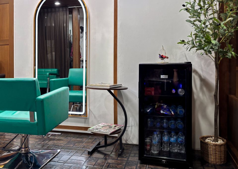 A teal styling chair before a tall arched mirror lit around its edge, with a round side table and an olive tree beside it.